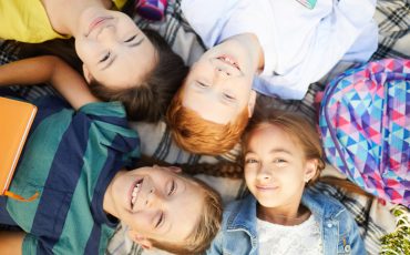 Four school friends lying on grass outside in park, smiling and enjoying end of last day of classes